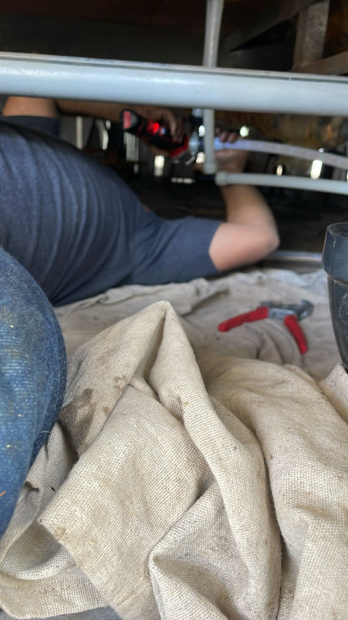 A person lying on their side under a sink, using a tool to work on gray plumbing pipes.