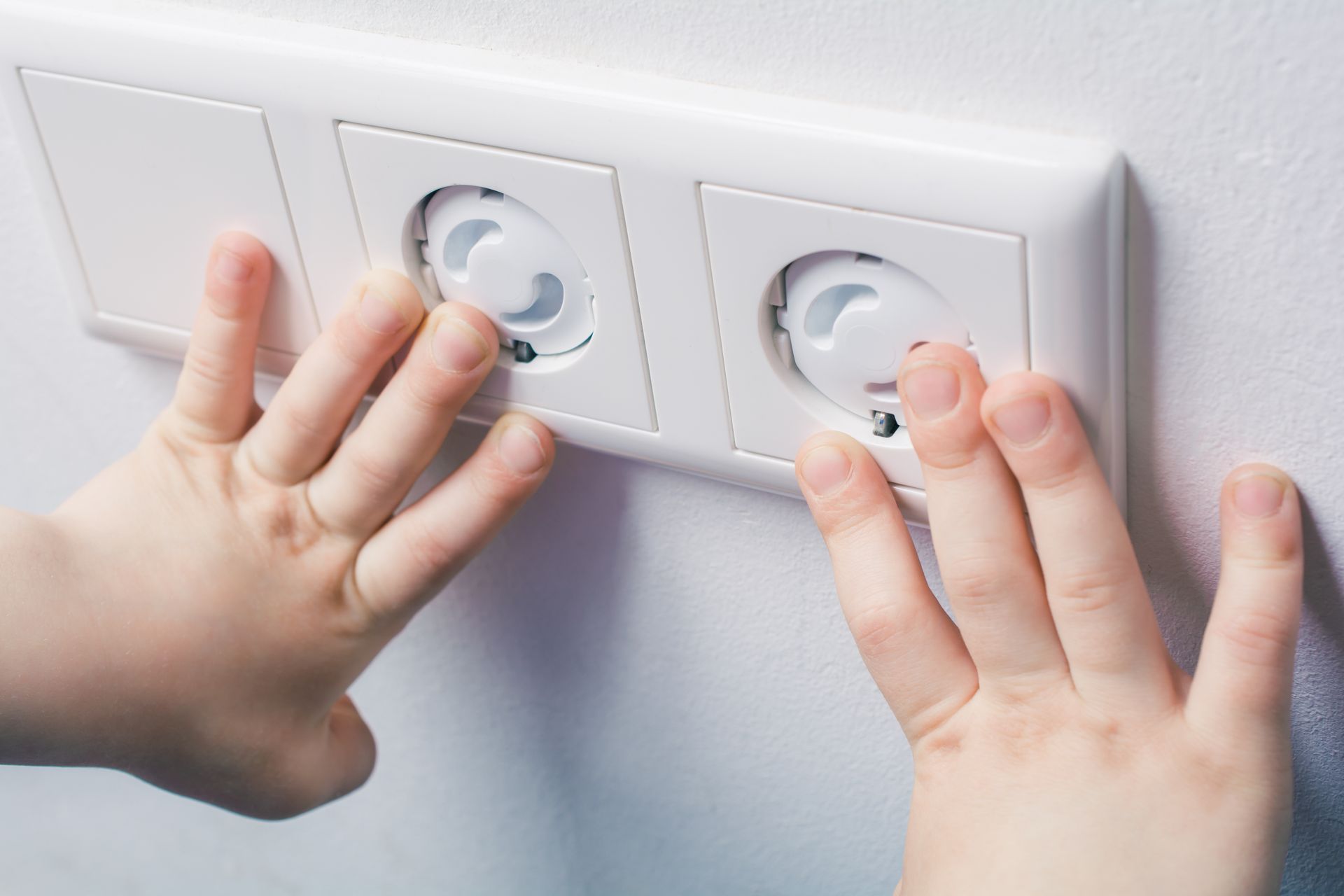 Two small hands reach out to touch a wall socket, but are protected by safety plugs, emphasizing the concept of preventing child hazards.