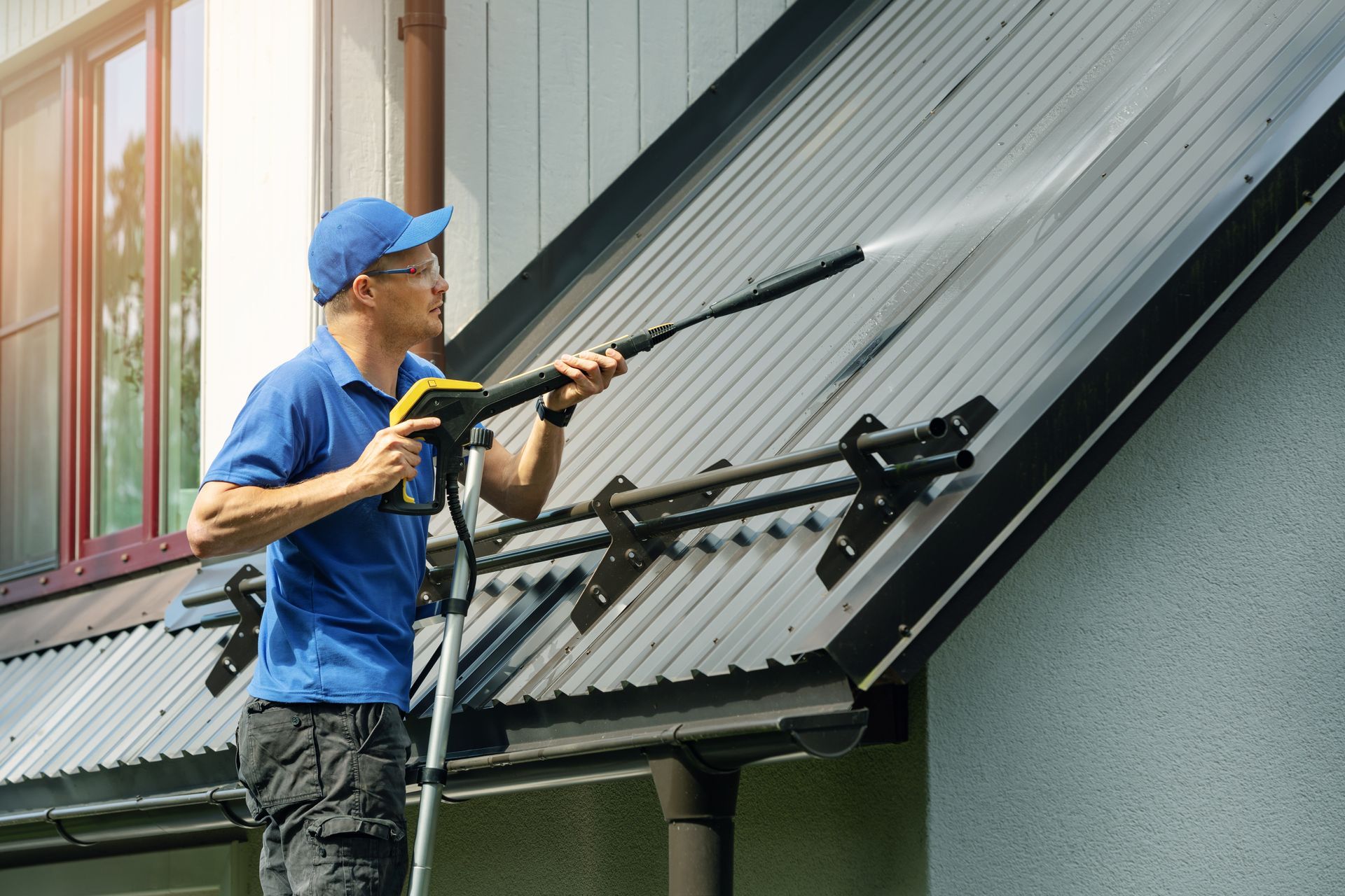 A man wearing protective gear stands on a ladder while using a high-pressure washer to clean a metal roof of a house.