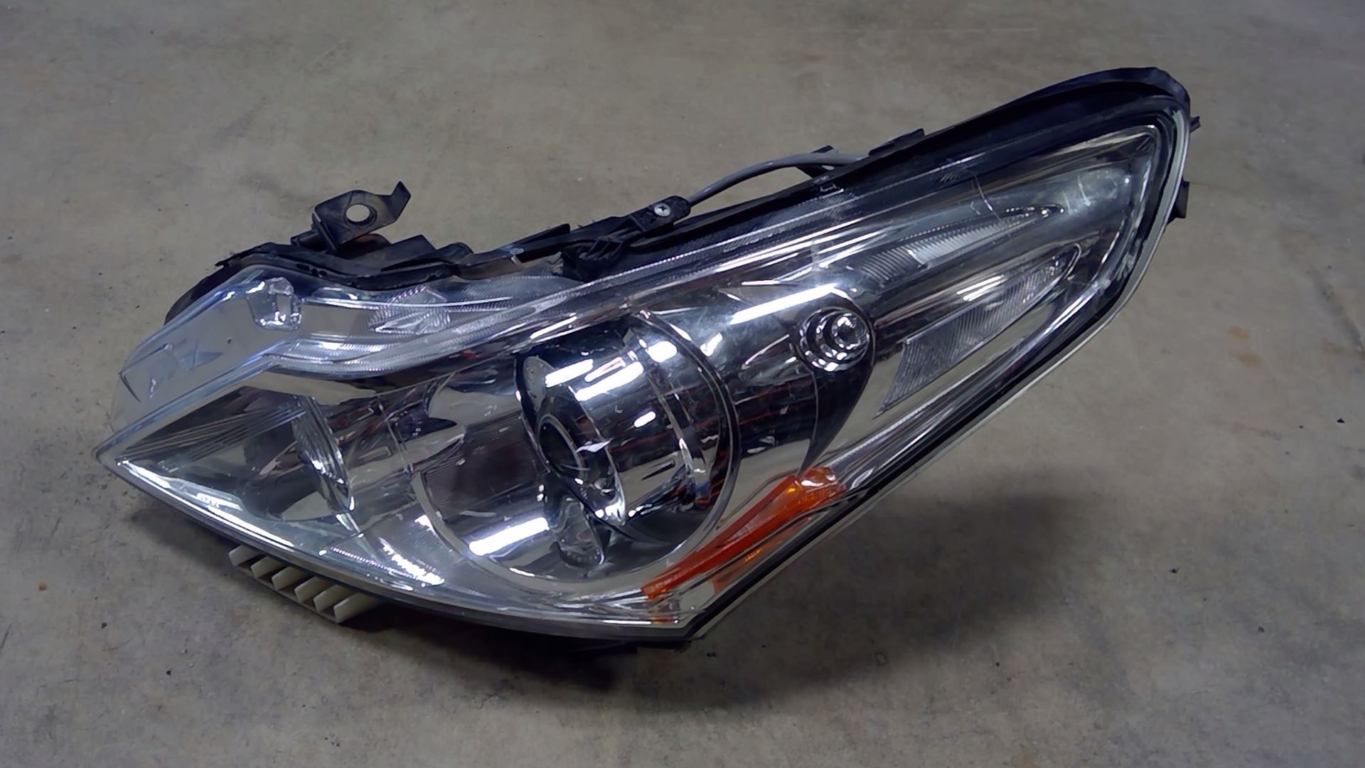 A single car headlight assembly with a clear lens and a small orange reflector, positioned against a neutral background.