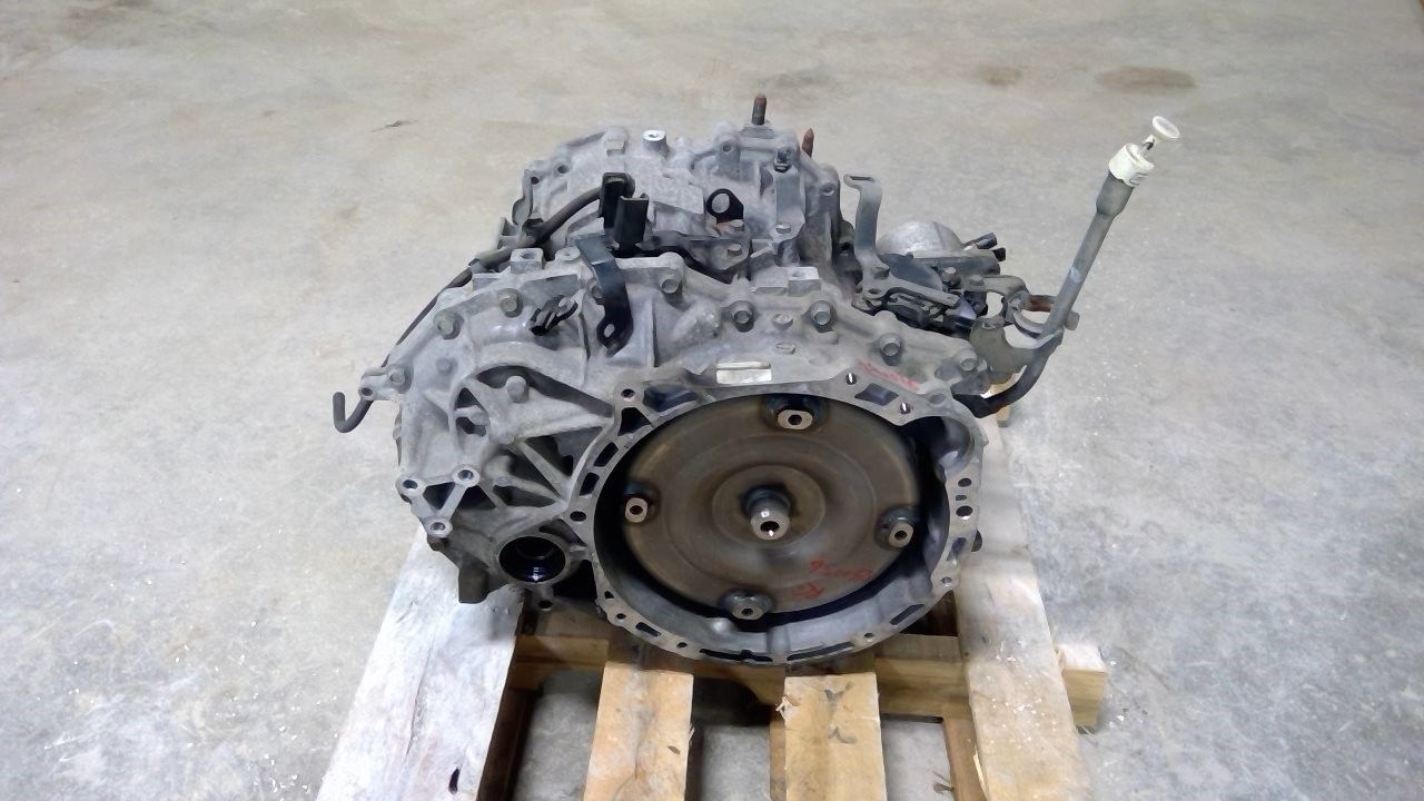 A metallic automotive transmission unit sits on a wooden pallet against a gray concrete floor background.