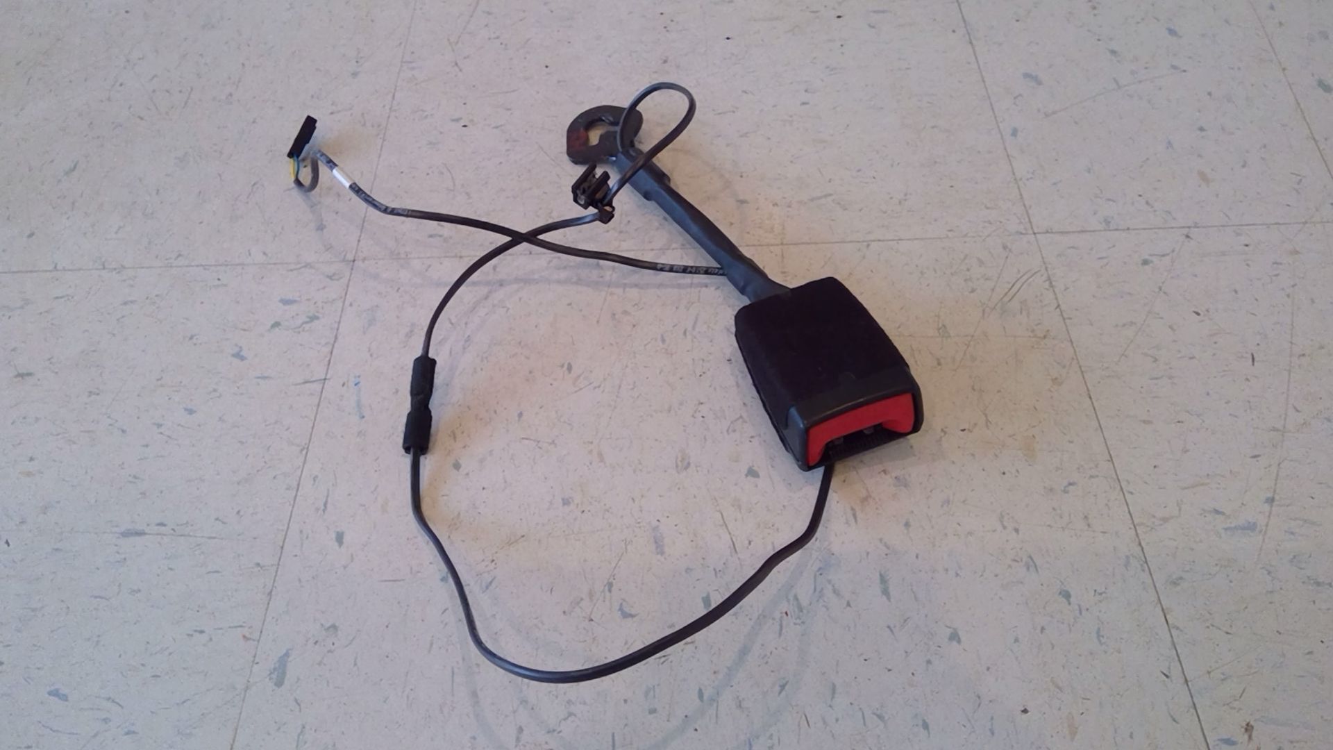 A car seat belt buckle with a mounting bracket and electrical wire connector, isolated against a white tiled background.