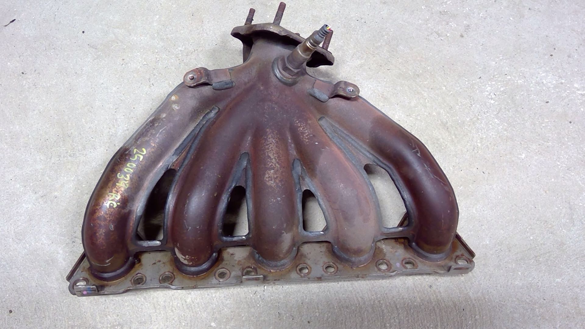 A rusty, four-runner cast-iron exhaust manifold with a threaded oxygen sensor port, viewed from the engine-mounting side.
