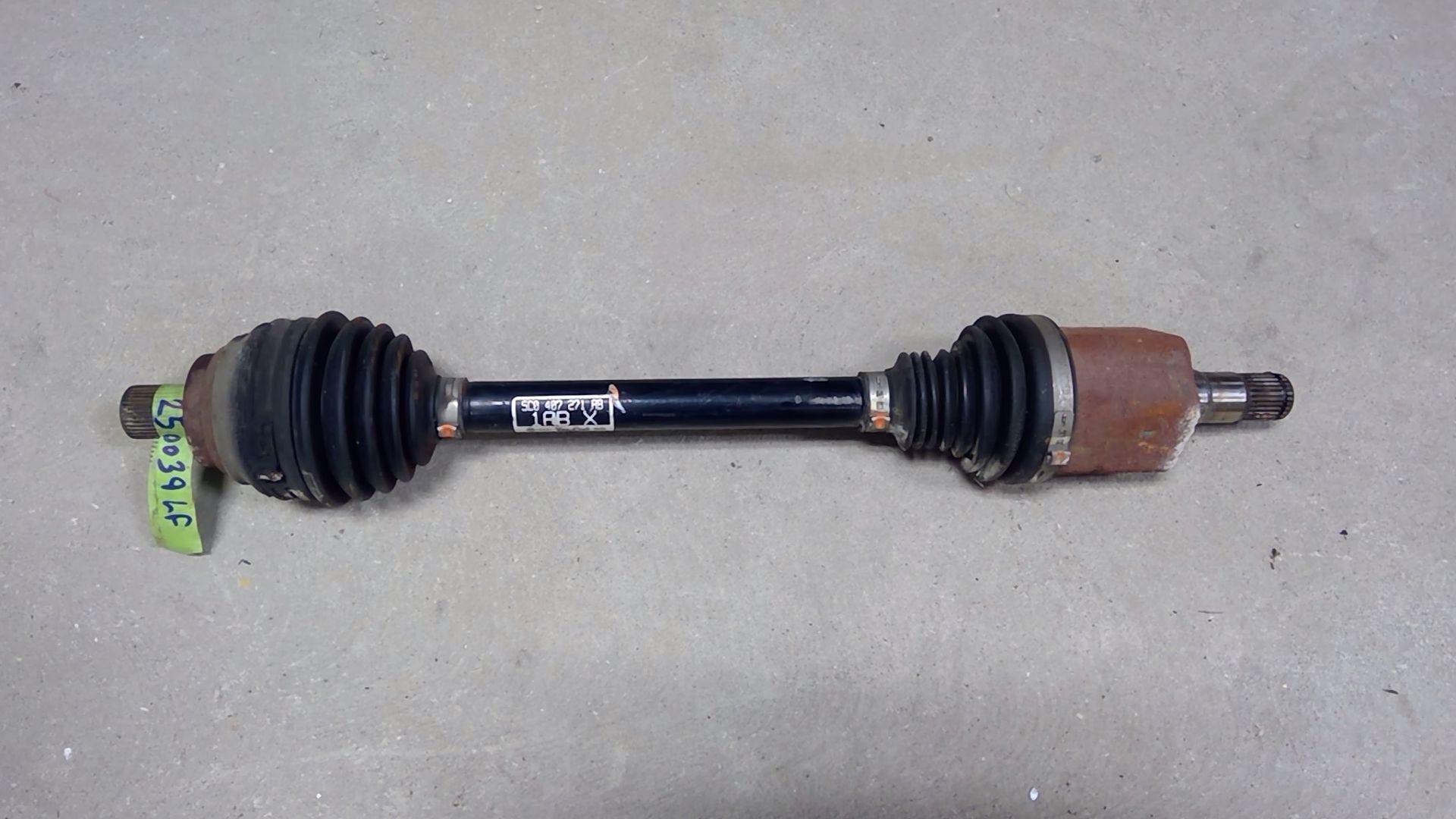 A black automotive CV axle shaft with black rubber boots and rusted metallic ends lying on a grey surface.