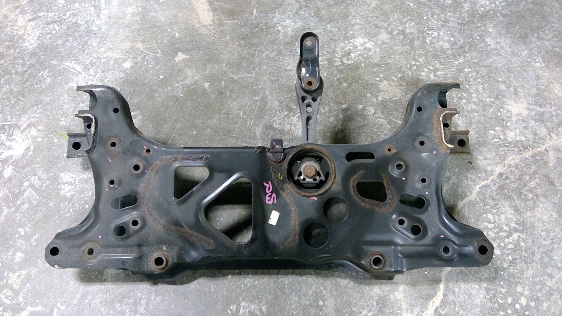 A black automotive subframe lying on a grey concrete floor, featuring mounting holes and a central engine mount bracket.