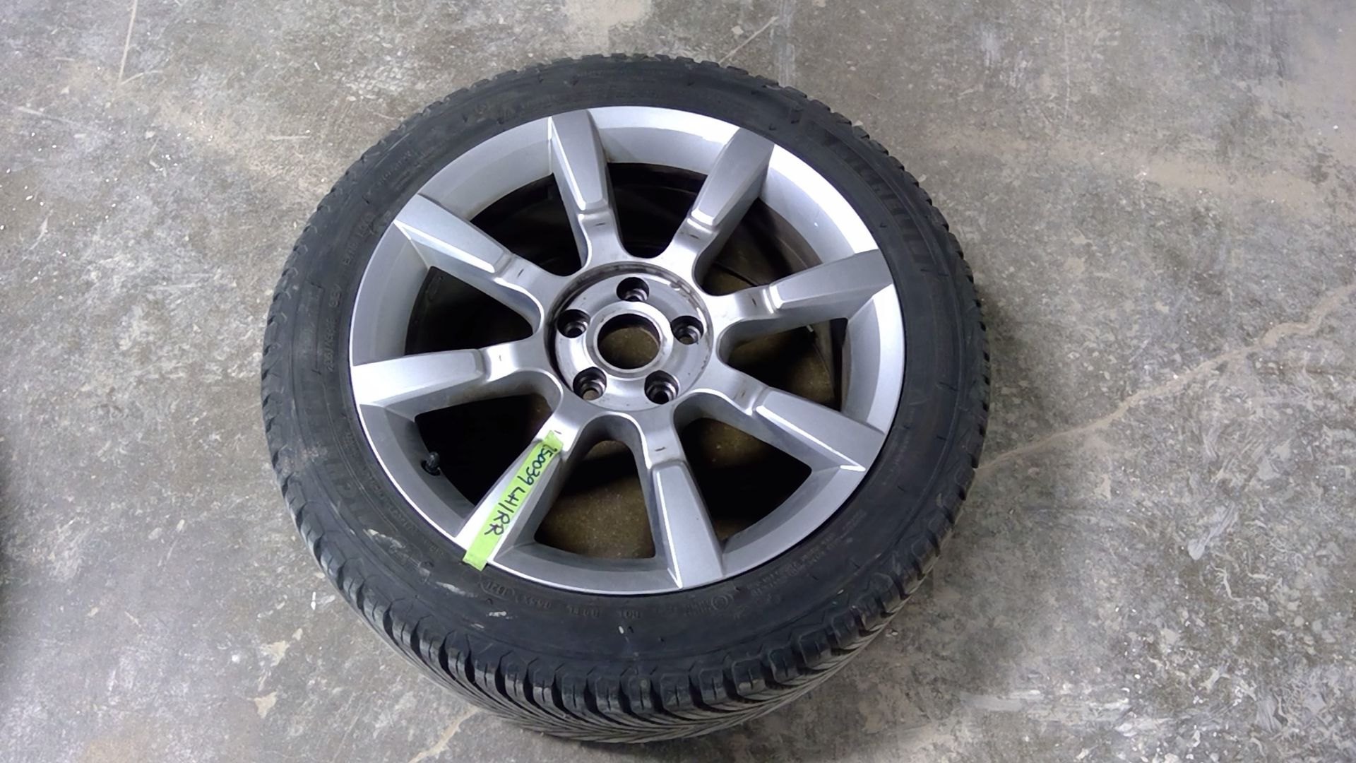 A silver, multi-spoke car wheel and tire on a grey concrete floor, featuring a small green tag attached to one spoke.