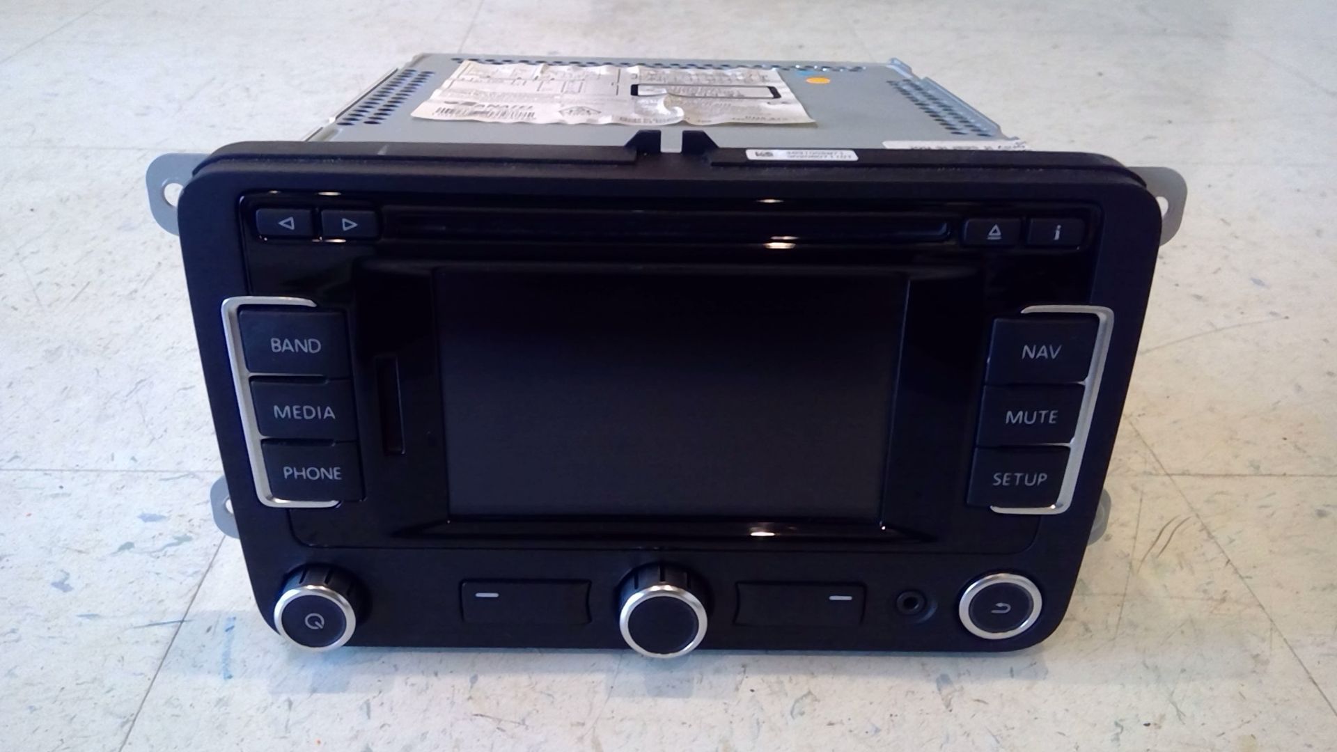 A black Volkswagen vehicle infotainment head unit with a central screen, physical buttons, and three circular knobs.