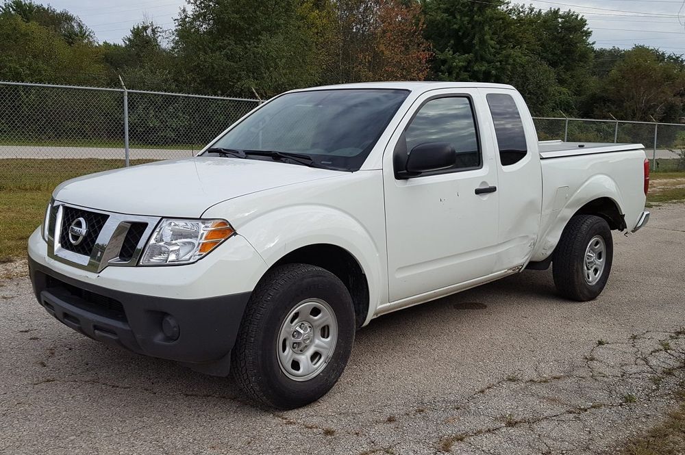 A white Nissan Frontier extended-cab pickup truck parked on a gravel lot with a chain-link fence in the background.