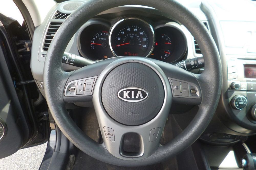 A black Kia steering wheel with mounted audio and cruise controls, positioned in front of the vehicle's dashboard gauges.