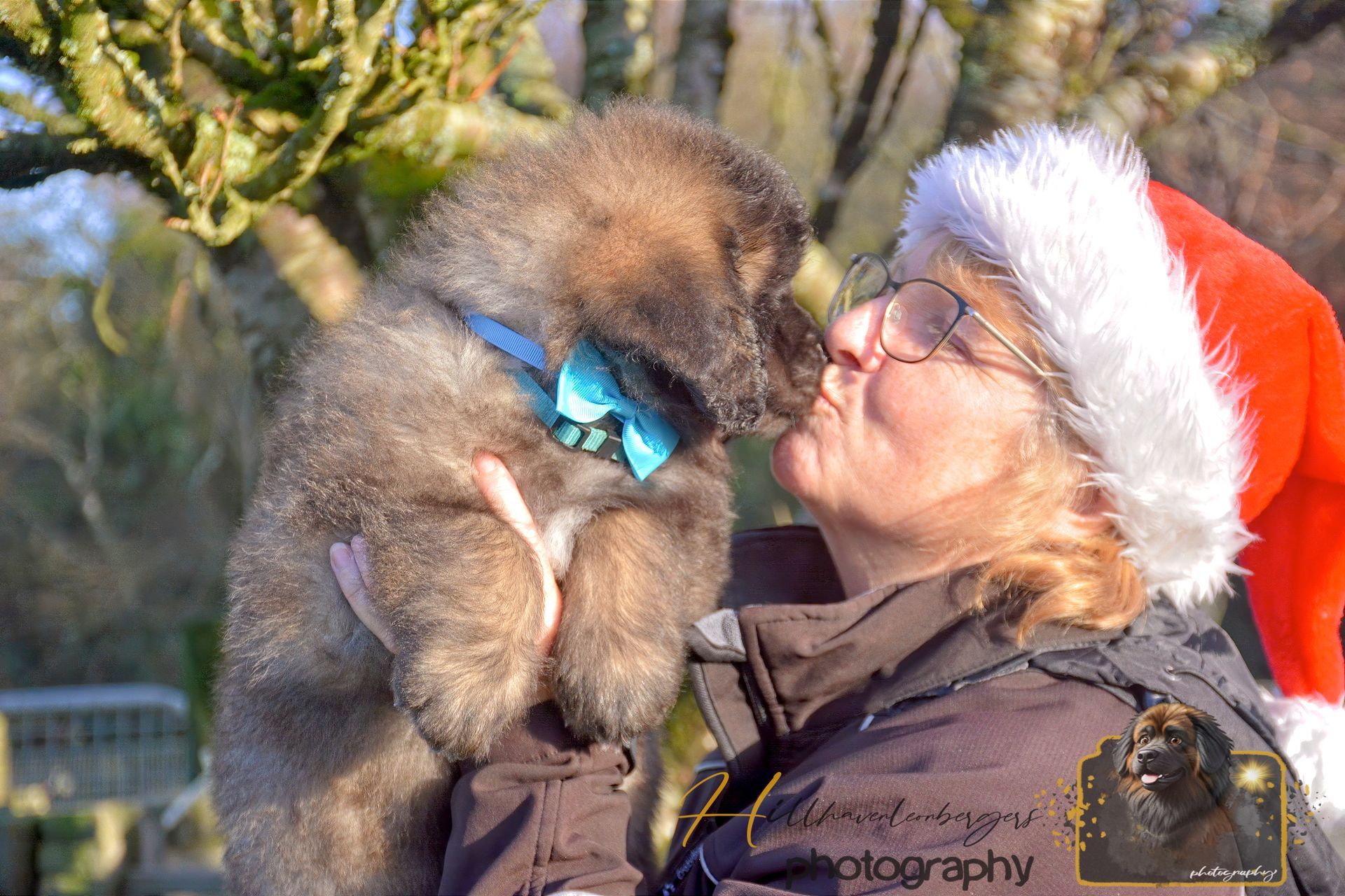 Woman in Santa hat kissing a fluffy puppy with blue bow tie outdoors.