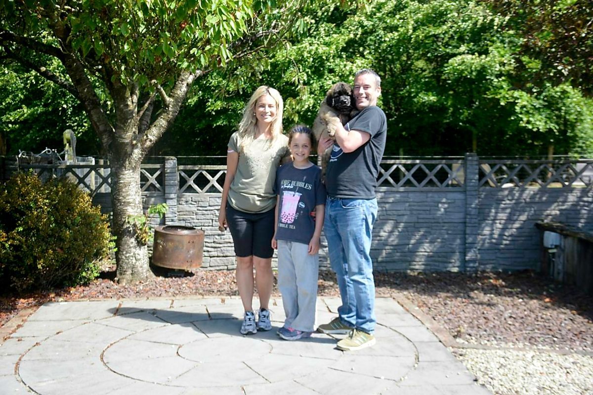 Family of four poses outdoors; man holding small black dog. Woman and child stand beside them, all smiling. Paved patio area with trees.