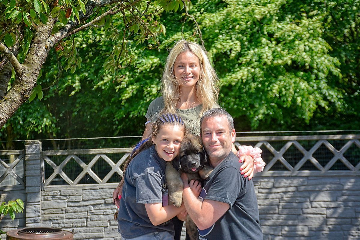 Family posing outdoors with a large, brown dog. They are smiling, embraced near a stone wall and green foliage.