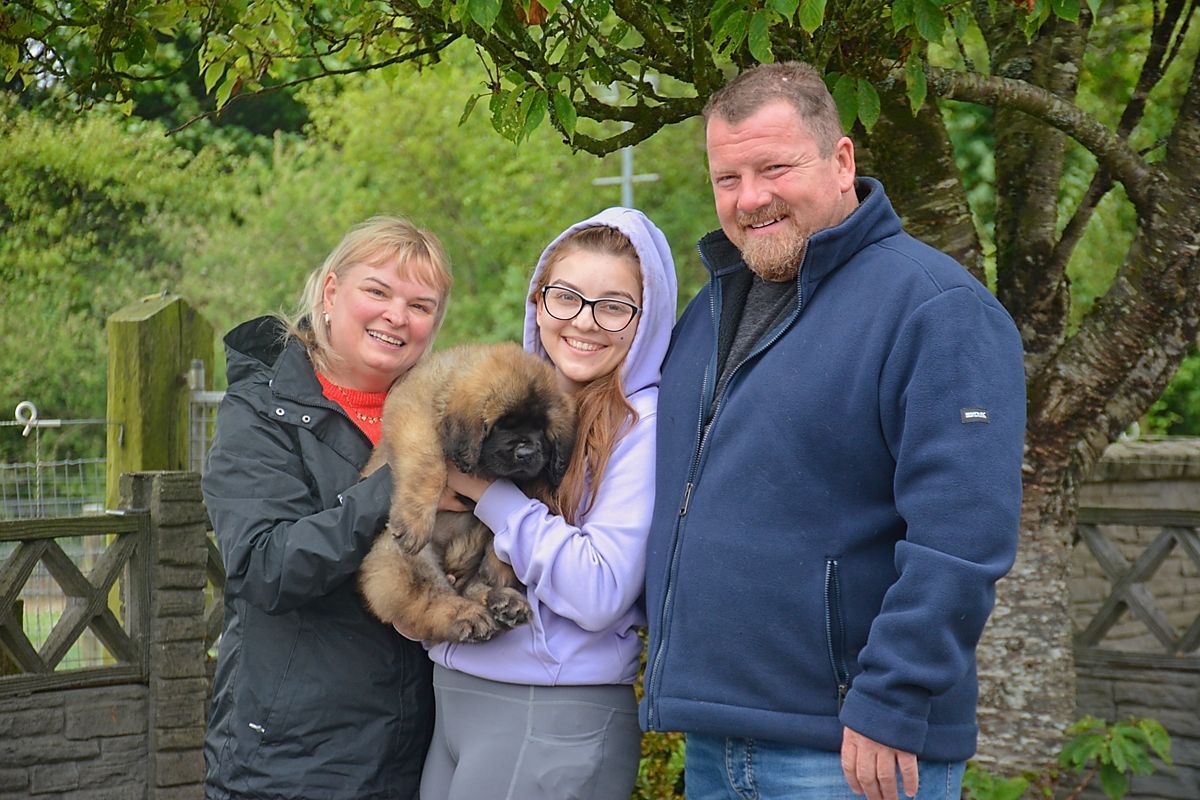 Family poses with a fluffy, brown puppy outdoors. The woman on the left holds it, while the others smile.