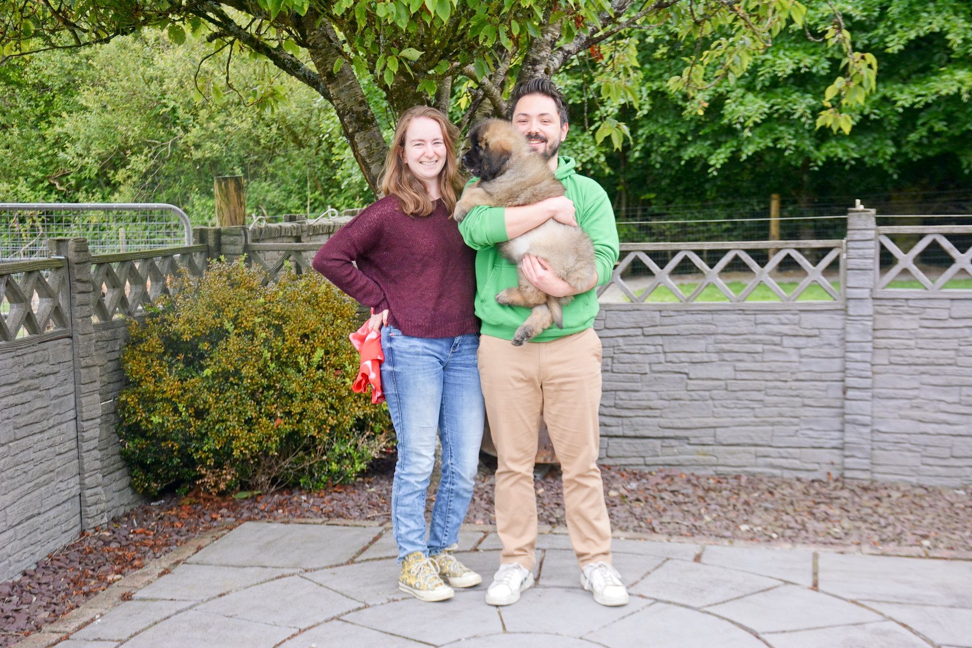 A couple posing outdoors, man holding a fluffy puppy, near a gray fence and greenery.