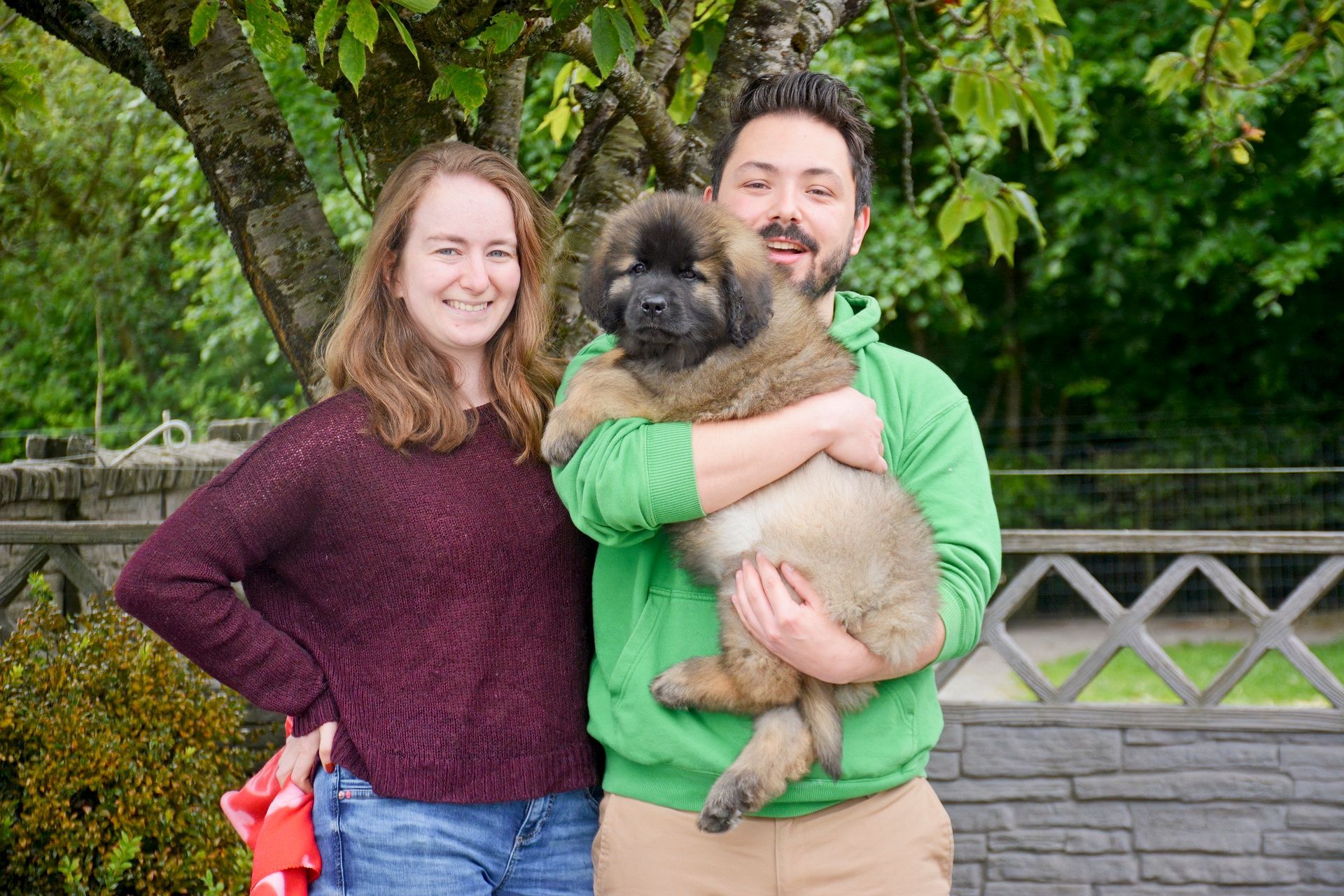 A couple holding a fluffy brown puppy outdoors. The man is smiling. The woman is smiling. Green hoodie.