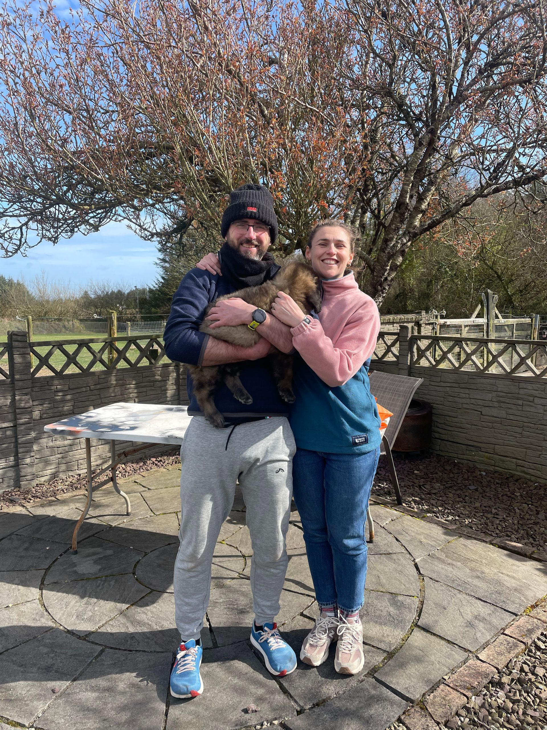 A couple standing on a patio holding a cat, with a large leafless tree and a fence in the background on a sunny day.