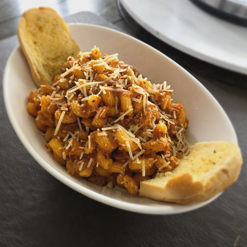 A bowl of pasta bolognese with garlic bread on a table