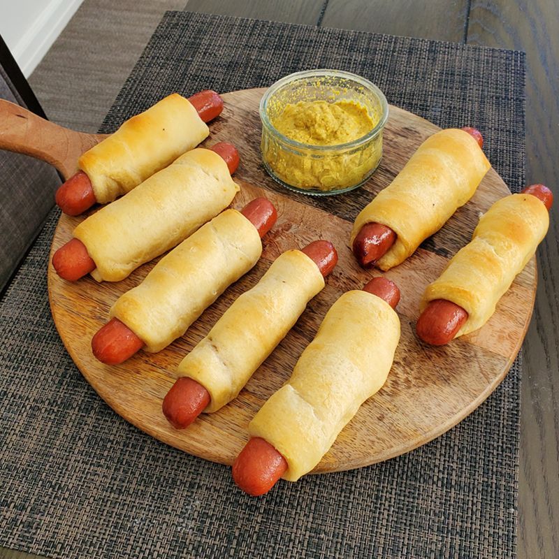 A wooden cutting board topped with hot dogs wrapped in dough and a bowl of mustard.