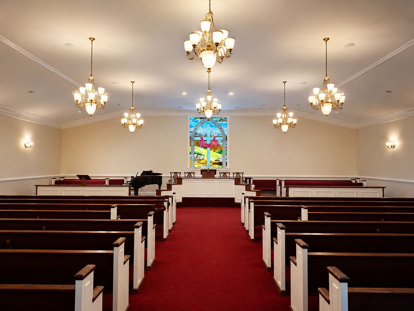 Empty church interior with red carpet, wooden pews, chandeliers, and a stained-glass window at the altar