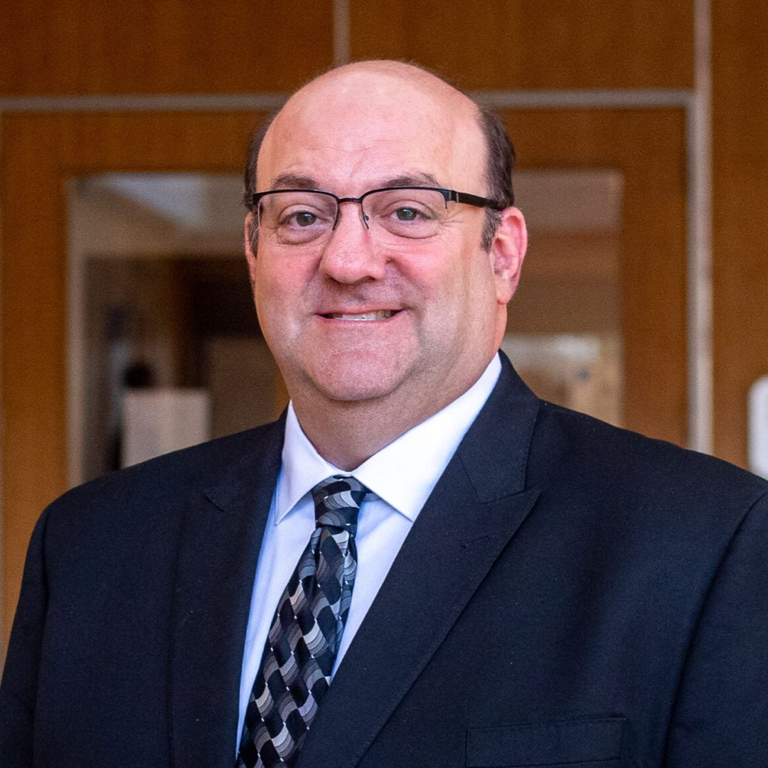 Man in black suit and glasses smiles, indoors.