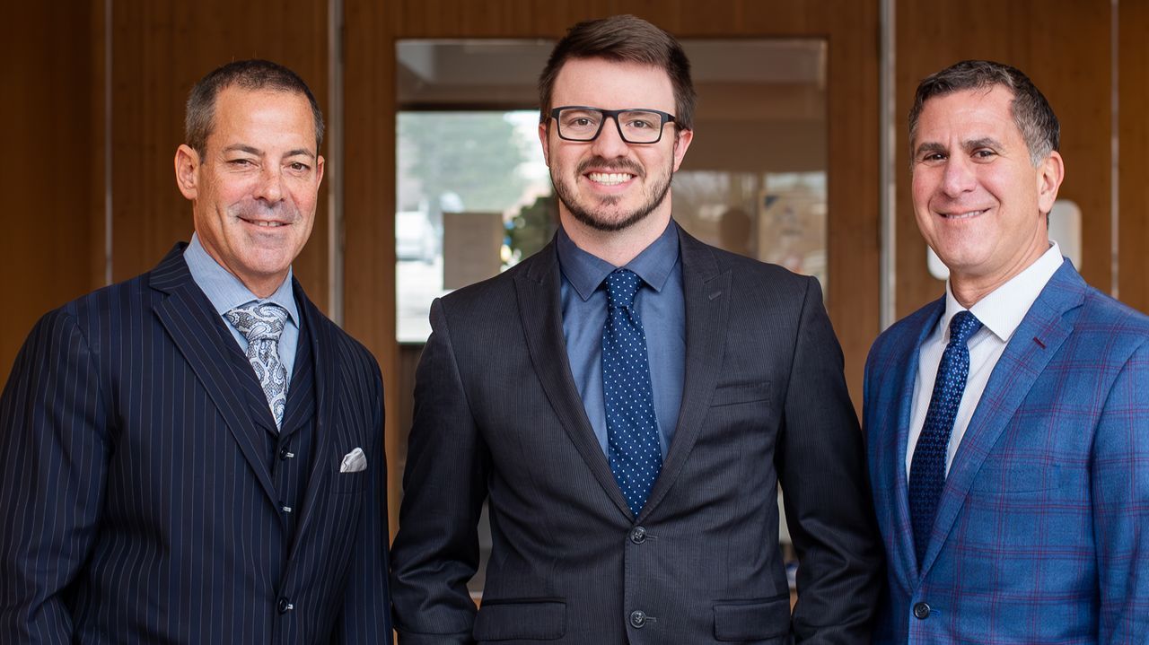 Three men in suits smiling, standing indoors, presumably a professional setting.
