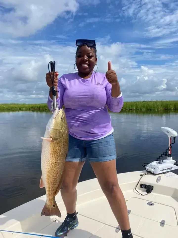 A woman is standing on a boat holding a large fish and giving a thumbs up.