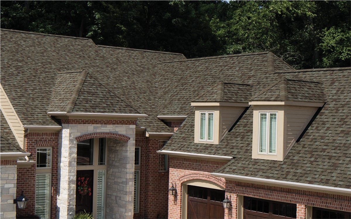 Close-up of a multi-gabled house with a brown shingle roof and dormer windows. The house features brick and stone exterior.