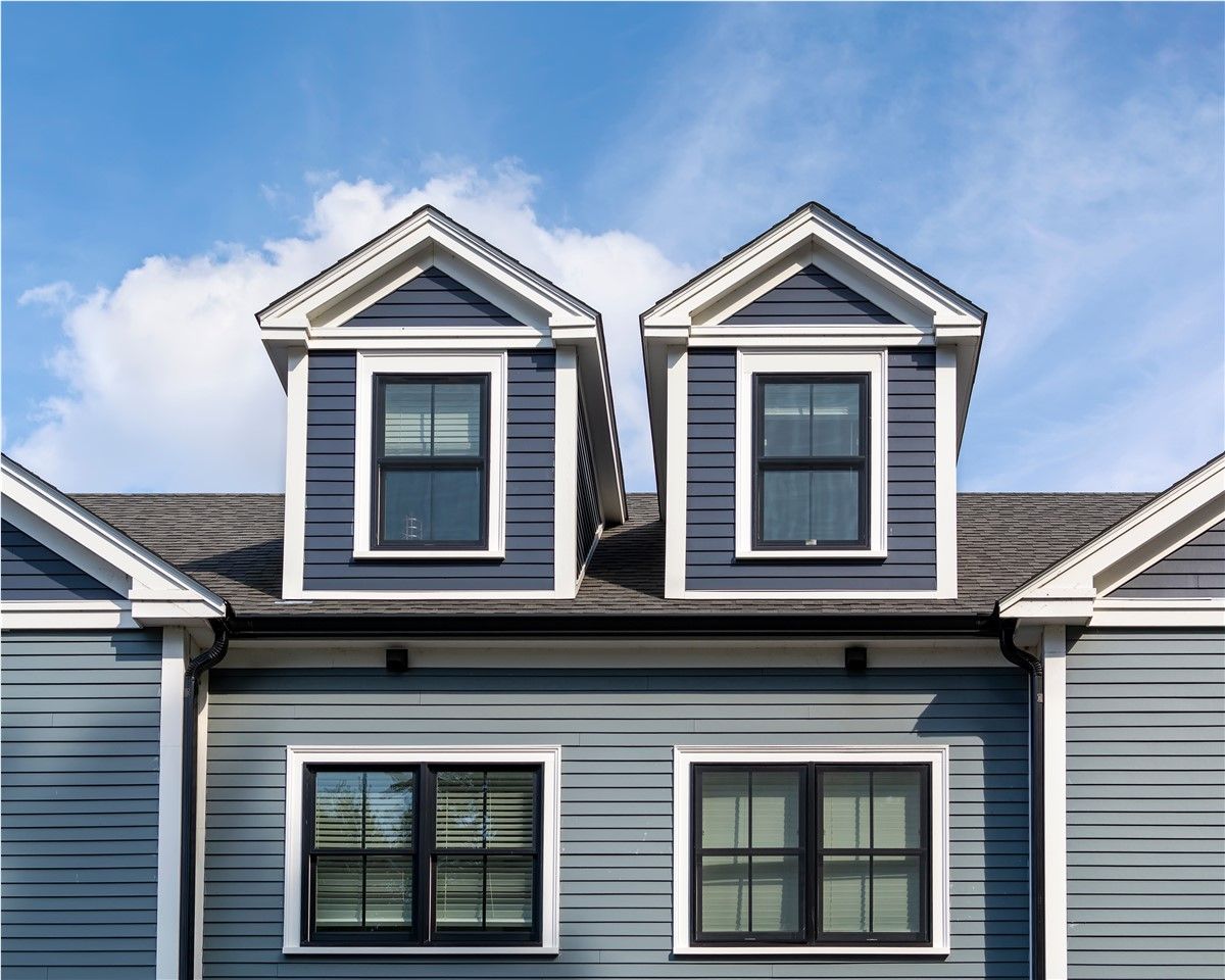 Blue and gray house with two dormers and windows against a partly cloudy sky.