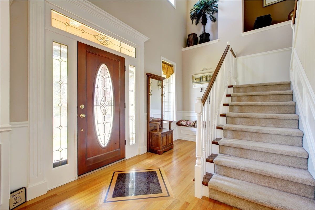 A bright entryway with a wooden door, sidelights, and a staircase with a neutral carpet, all set on a hardwood floor.