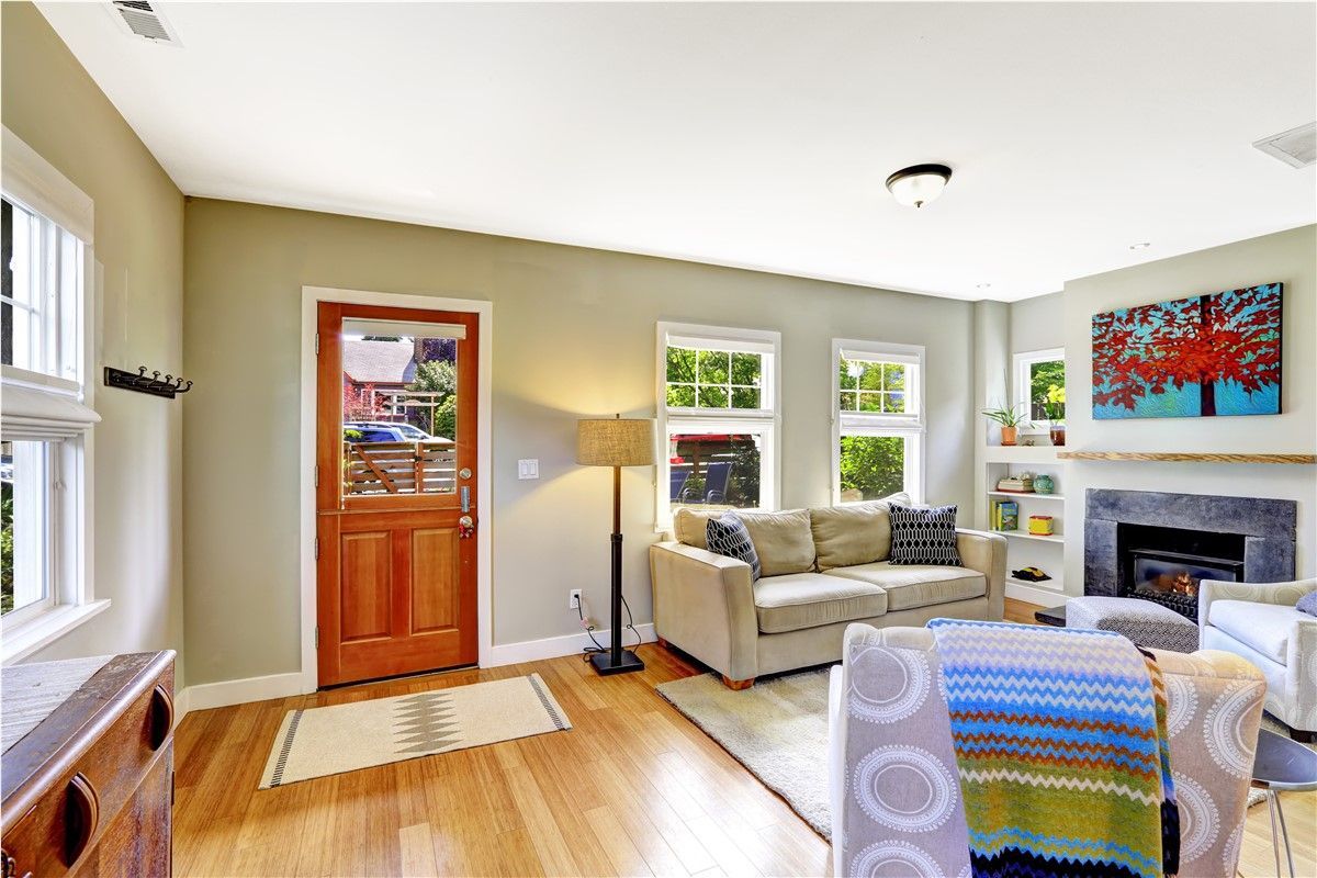 A bright living room with wooden floors, beige walls, and a wooden door. A tan couch and fireplace are visible.