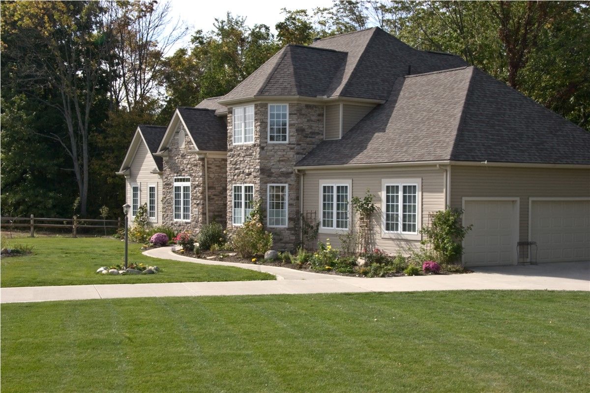 Two-story house with stone and tan siding, light roof, and two-car garage; front lawn with flowers and trees.