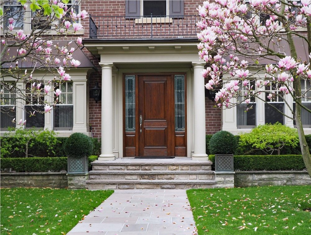 A brick home's front entrance with a wooden door, flanked by columns and blooming pink trees.