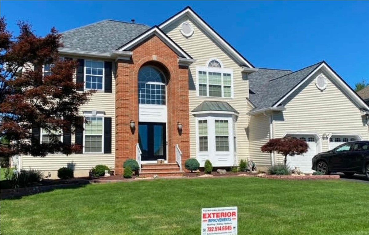 Two-story house with beige siding, a brick accent, and a new gray roof on a sunny day; a car is parked in the driveway.