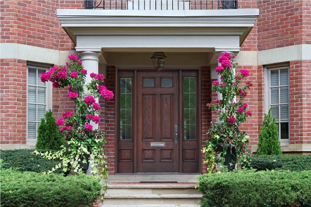 Red brick house entrance with dark wooden door and vibrant pink flowers climbing pillars.