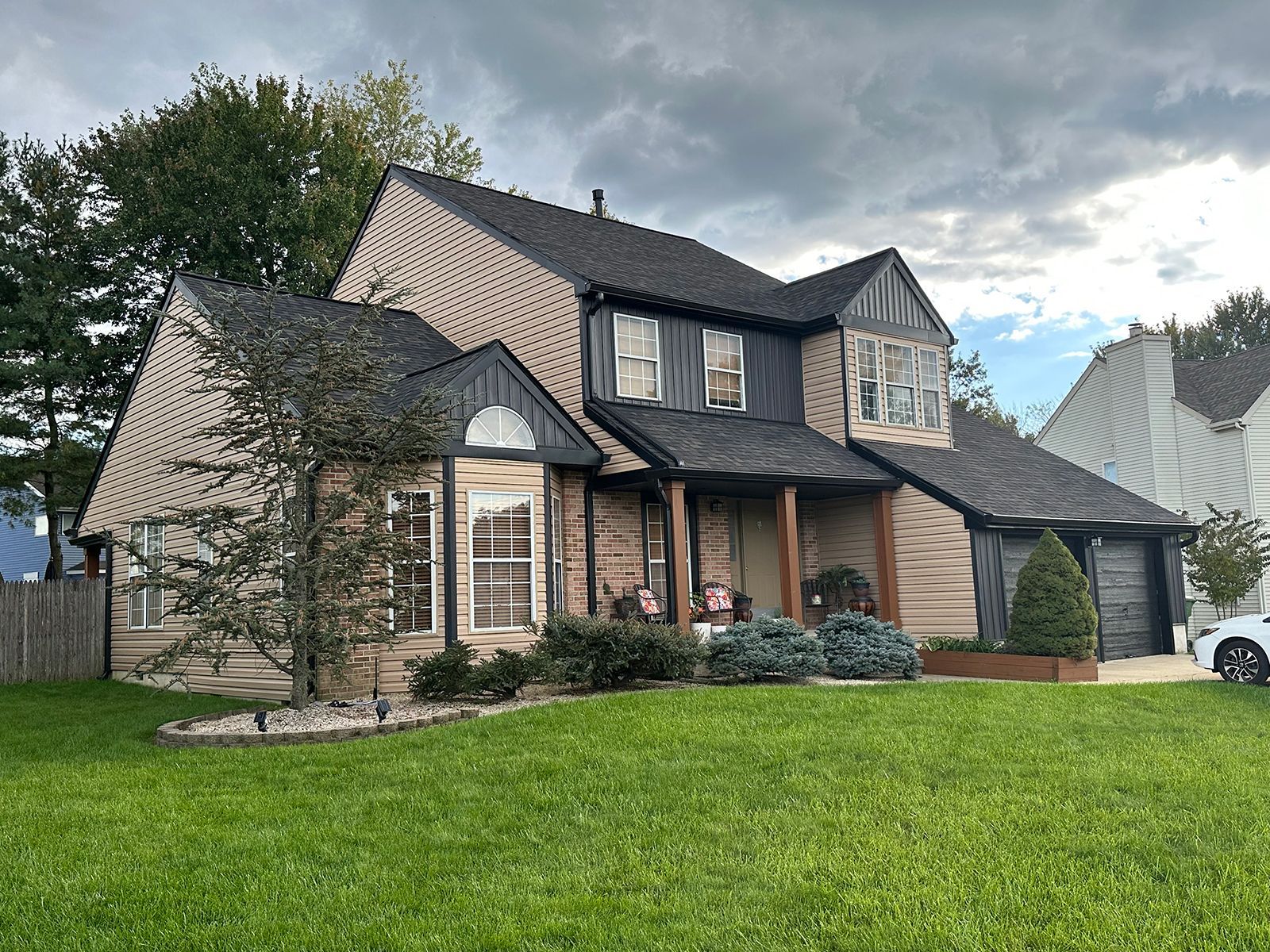 Two-story beige house with black accents, green lawn, and a cloudy sky in the background.