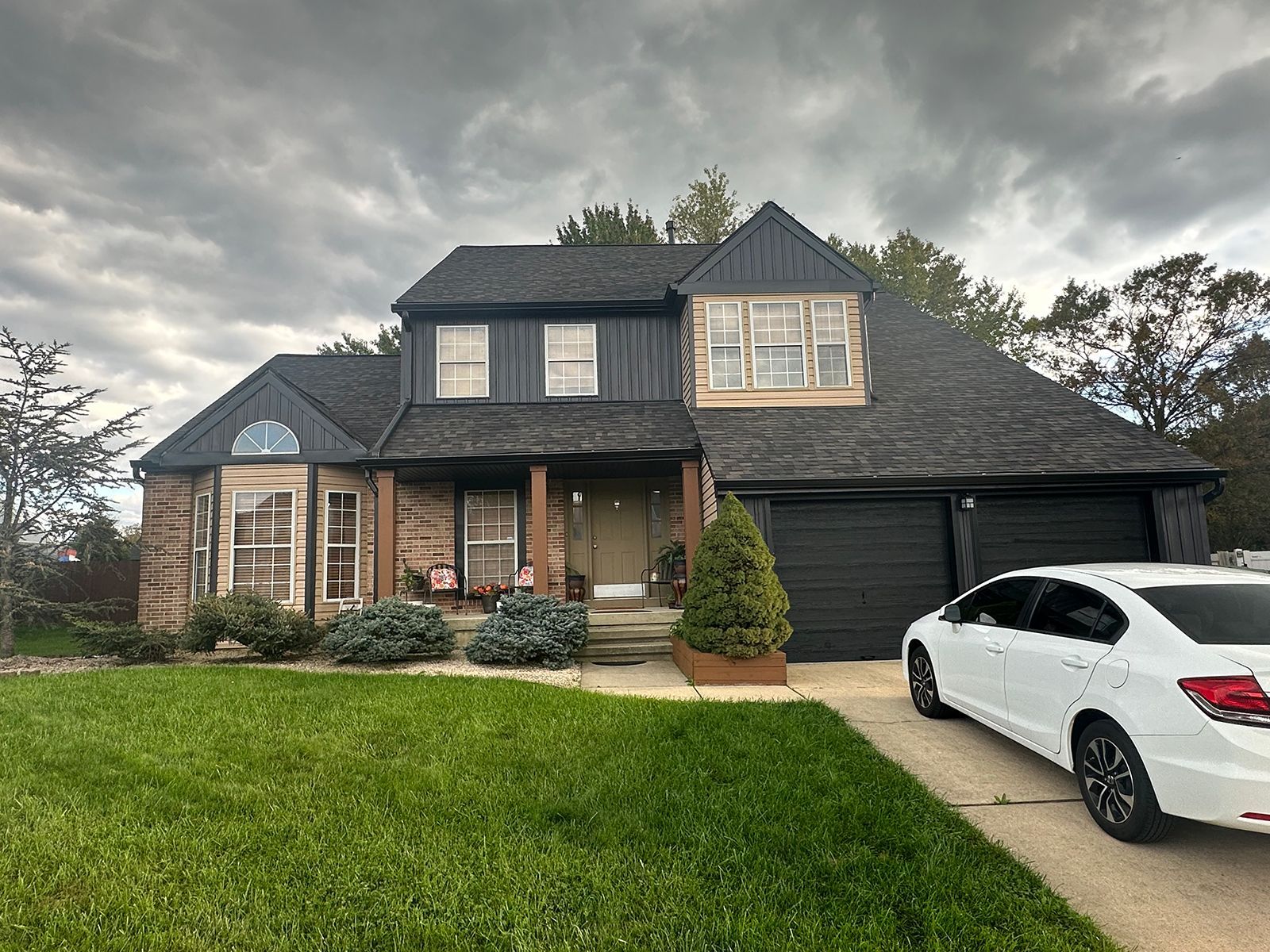 A two-story house with black trim and a black garage door. A white car sits in the driveway on a cloudy day.