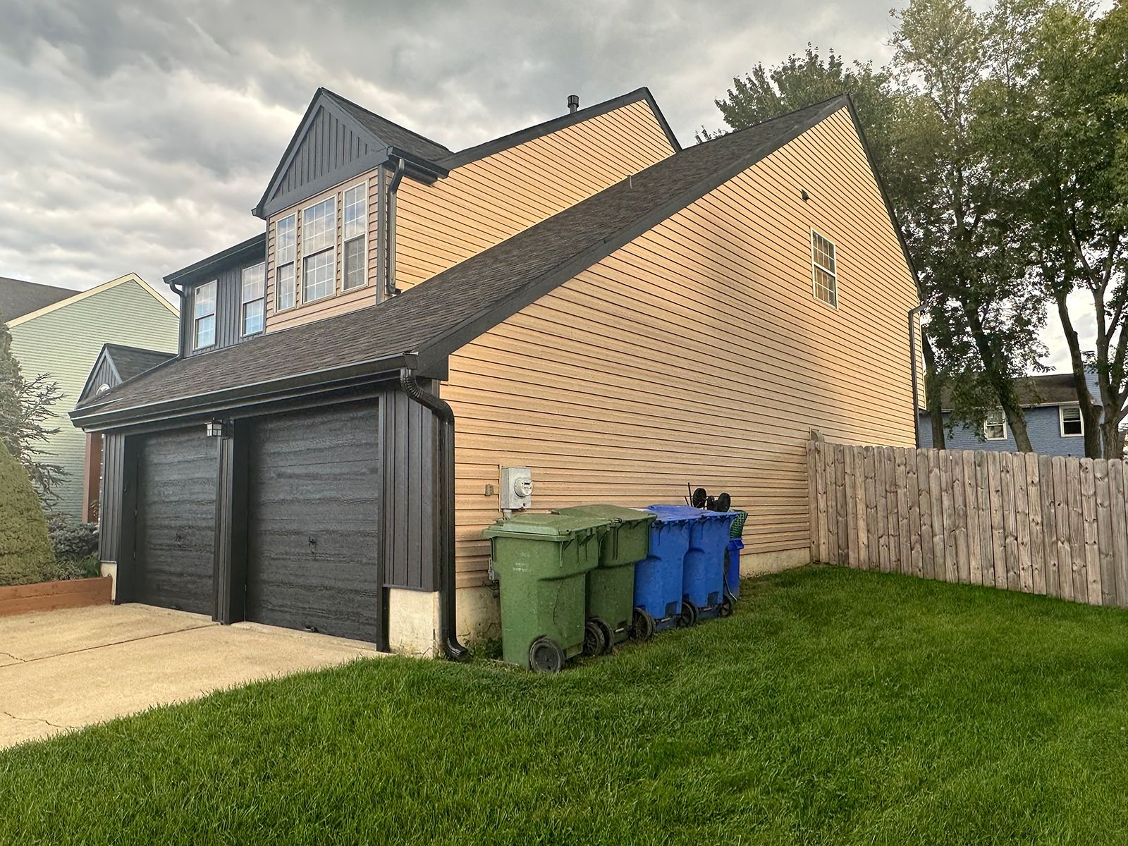 Two-story house with black garage doors, tan siding, and a row of trash bins on the lawn.