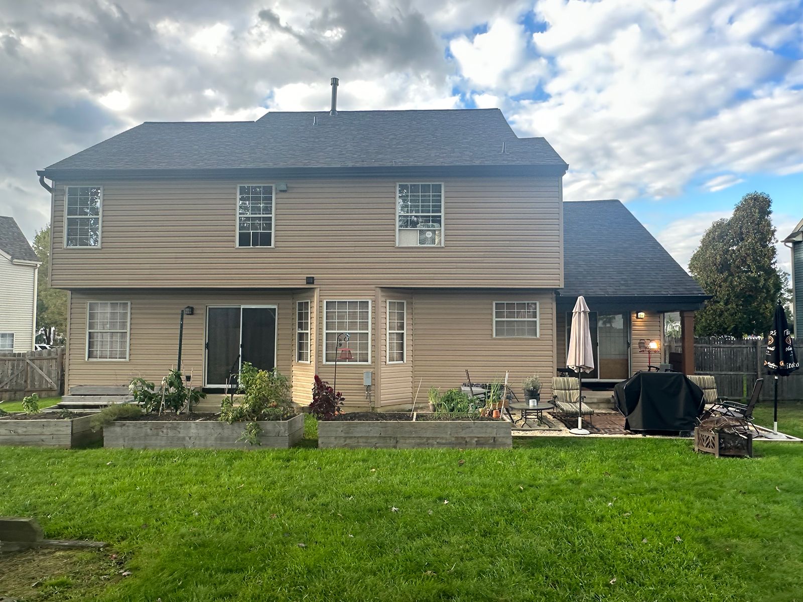 Backyard of a two-story house with tan siding, a dark roof, and a green lawn. A sliding glass door and windows are visible.