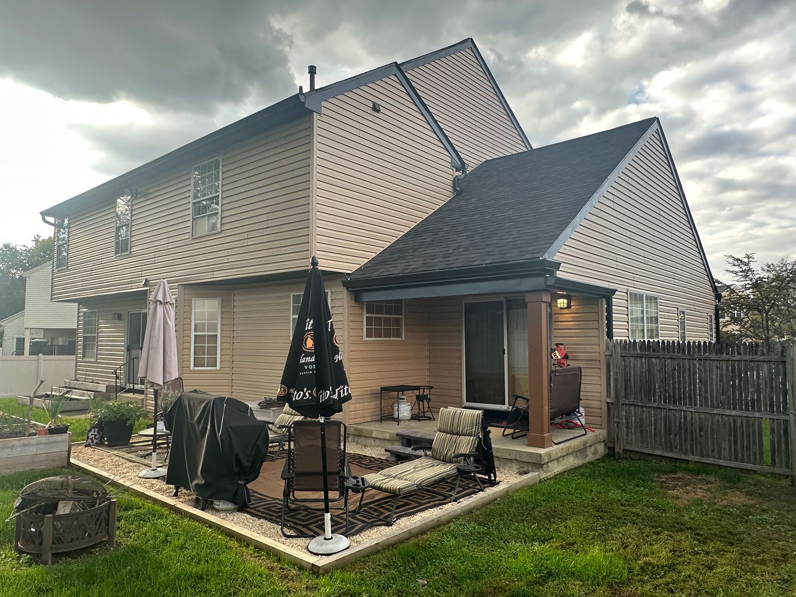 Backyard view of a two-story beige house with a patio, lawn, and wooden fence under an overcast sky.