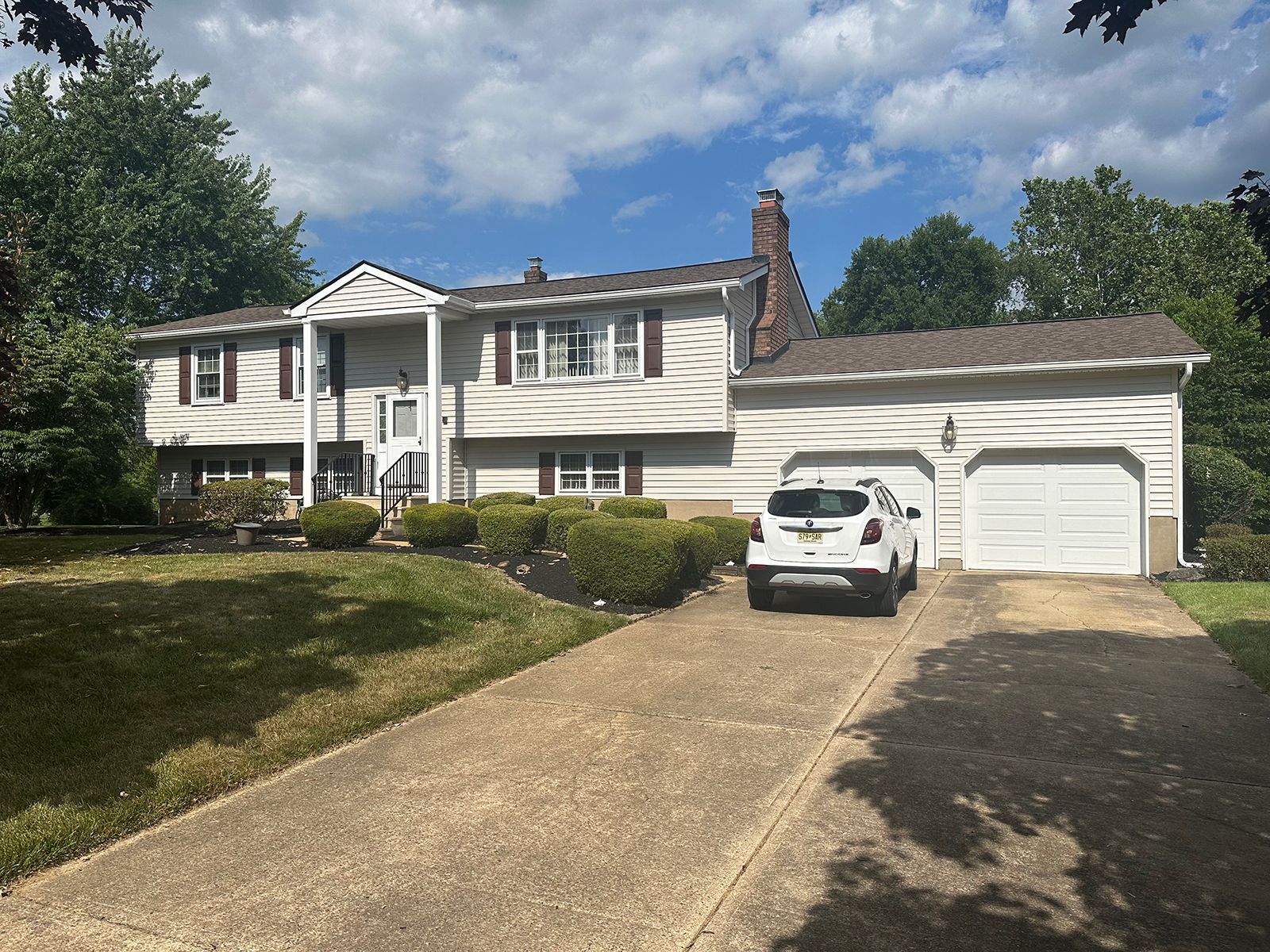 A two-story beige house with a two-car garage and a long driveway on a sunny day. A car is parked in front of the garage.