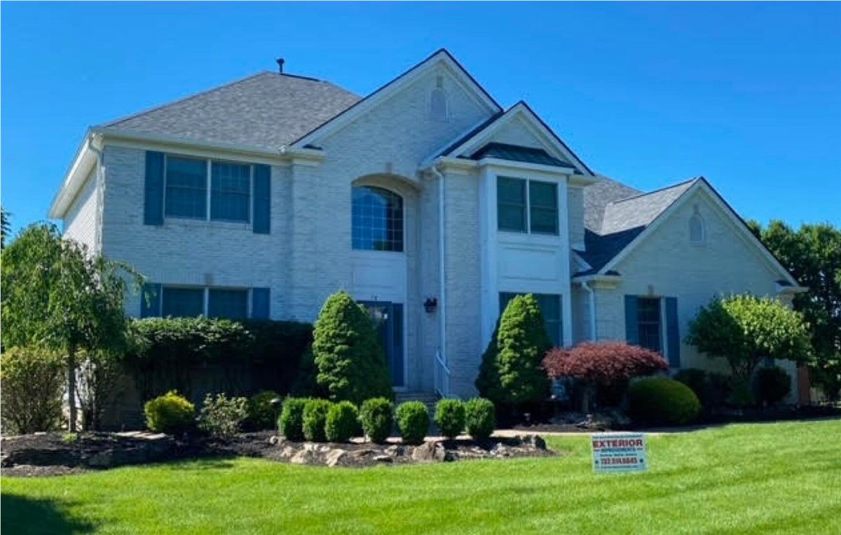Two-story light brick house with a dark roof, blue shutters, and well-maintained lawn under a clear blue sky.