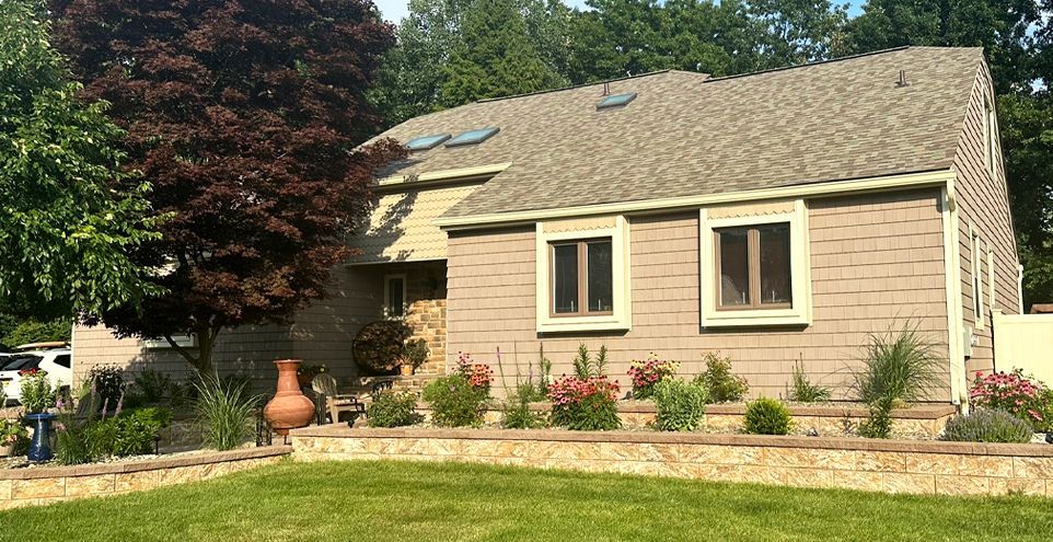 House with beige siding, brown roof, and tiered landscaping with a lush green lawn. Red and green trees frame the home.