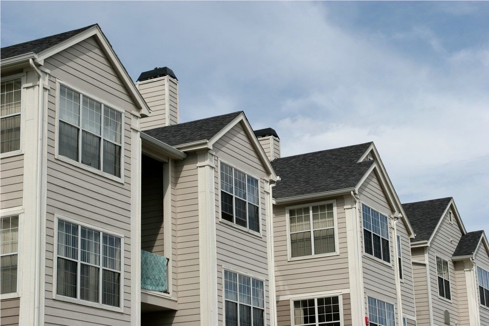 Row of beige apartment buildings with gray roofs and white trim against a blue sky.