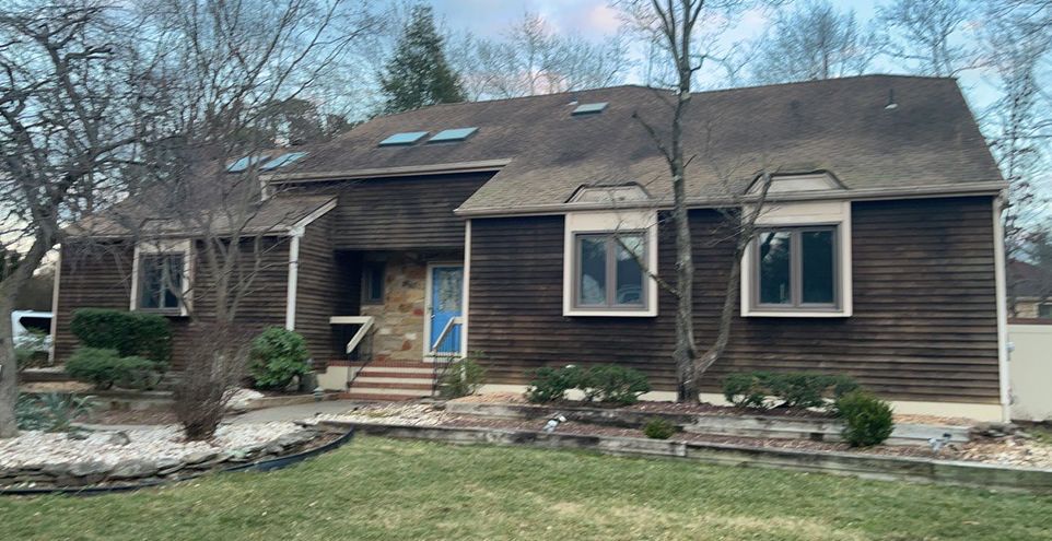 A one-story brown house with several windows, a blue front door, and a dark roof set amongst bare trees and shrubs.