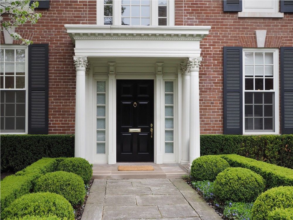 Brick house entrance with black door, white columns, and manicured landscaping. Black shutters flank windows on either side.