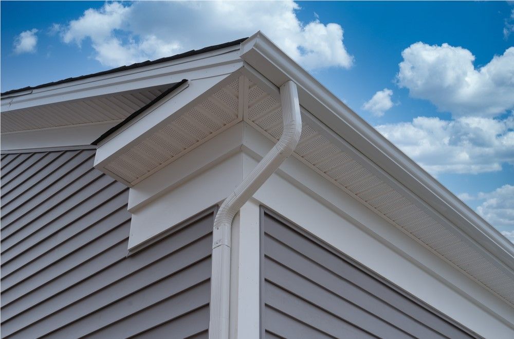Corner of a house with grey siding and white trim, including gutters and a downspout against a blue sky with clouds.