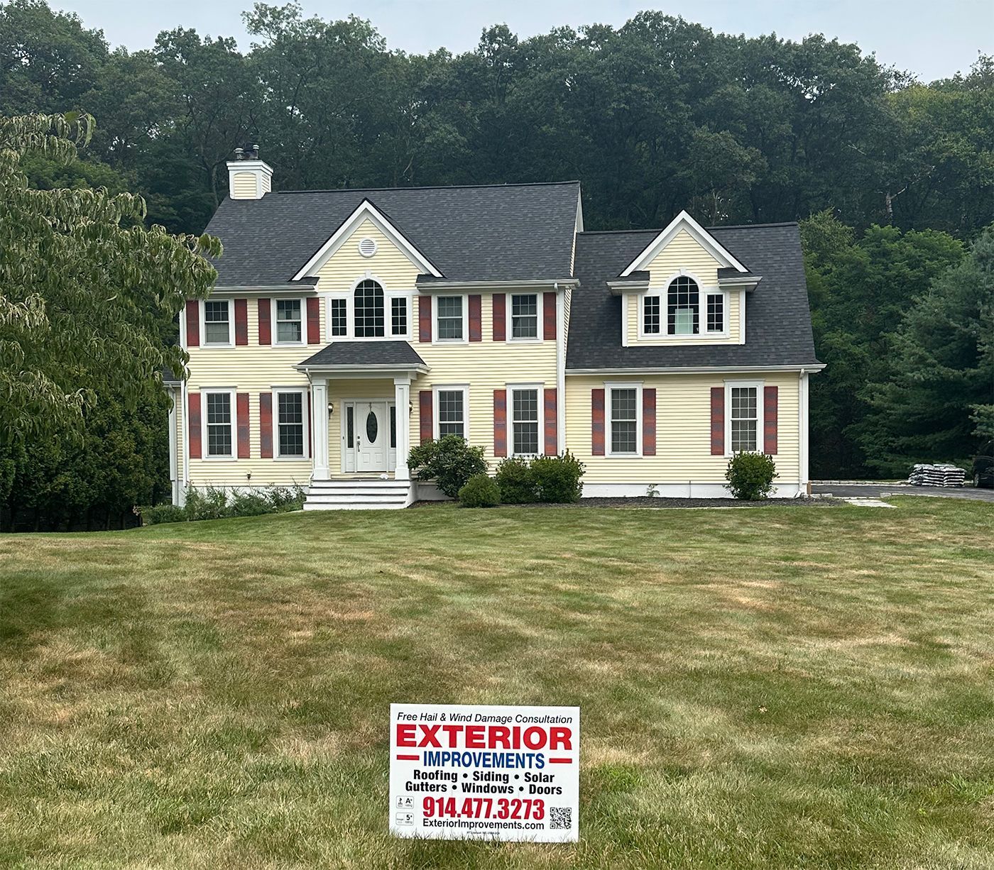 Two-story yellow house with dark roof, red shutters, white trim, on a grassy lawn. Exterior Improvements sign.