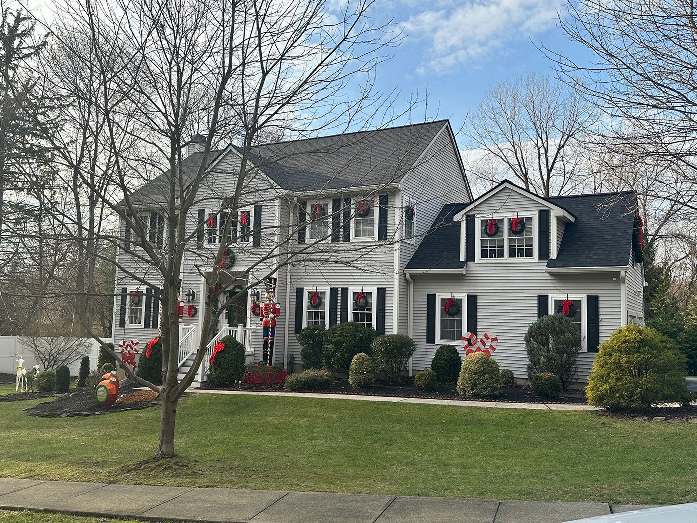 Two-story gray house with black shutters and wreaths, a green lawn, and bare trees on a cloudy day.