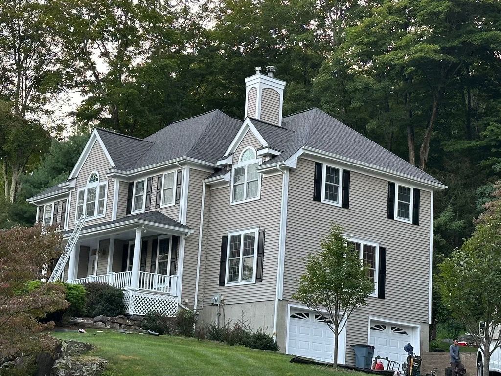 Two-story house with angled siding, black shutters, and grey roof, set on a hill surrounded by trees.