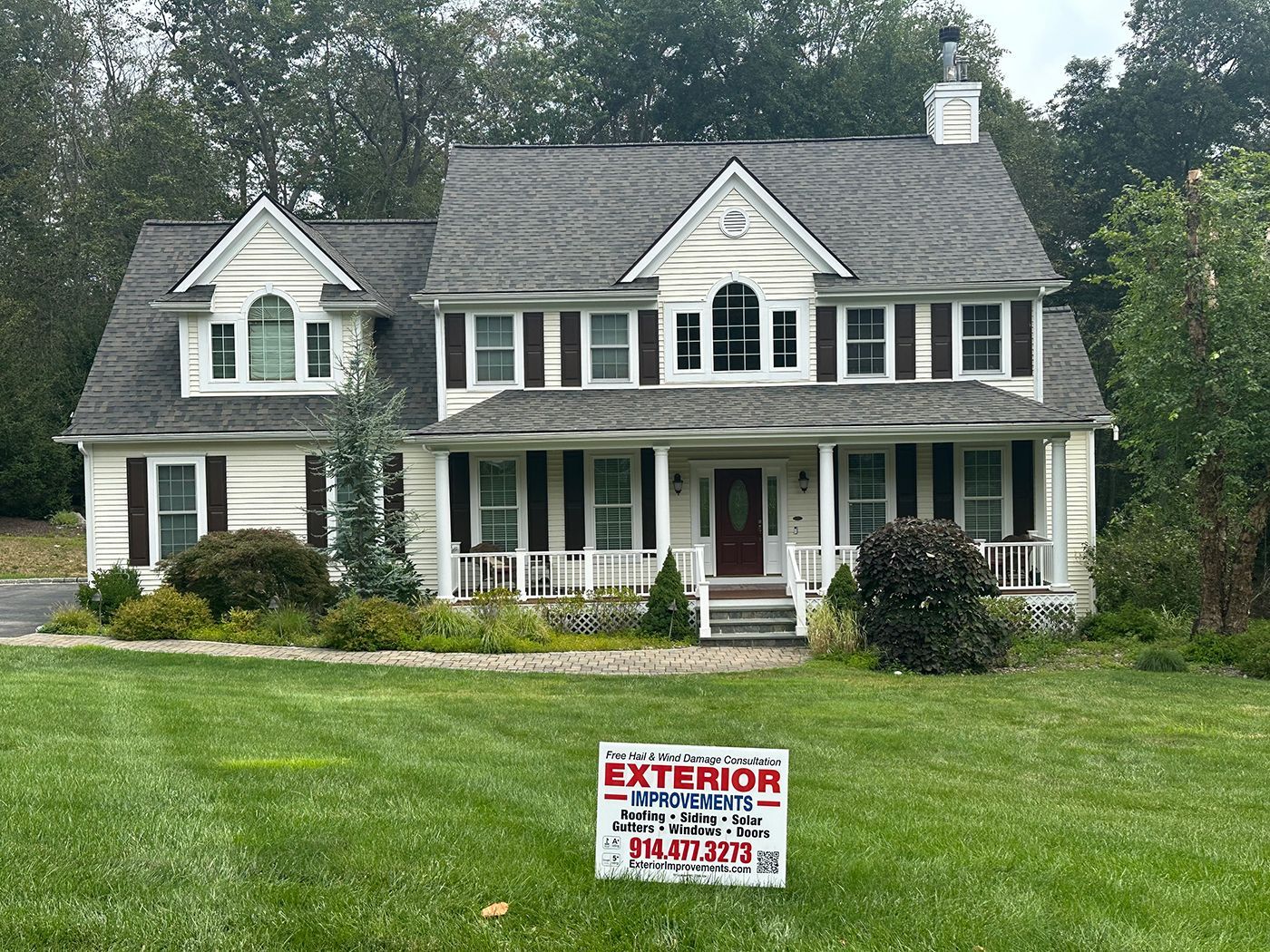 Two-story beige house with gray roof, black shutters, and sign on lawn.